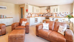 A modern kitchen with white cabinets, a tiled backsplash, and a central island. There are leather sofas and ottomans in the living area. - Crab Corner (Wells-next-the-Sea)