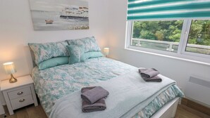 Een slaapkamer met een bed, kussens, een nachtkastje, een lamp, een raam met uitzicht en een schilderij aan de muur.