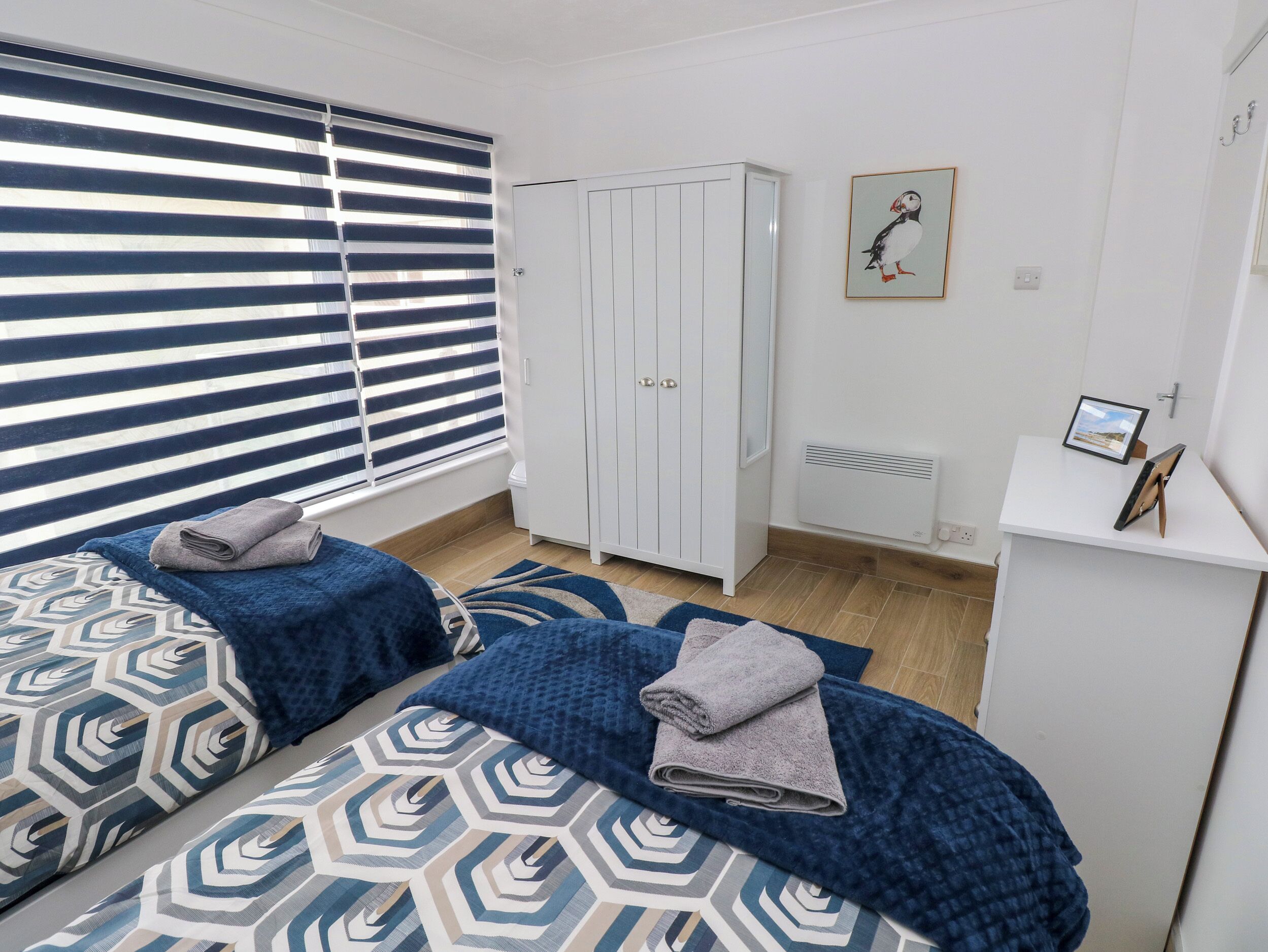 A room with two beds, a wardrobe, a radiator, and a small desk with a picture frame.