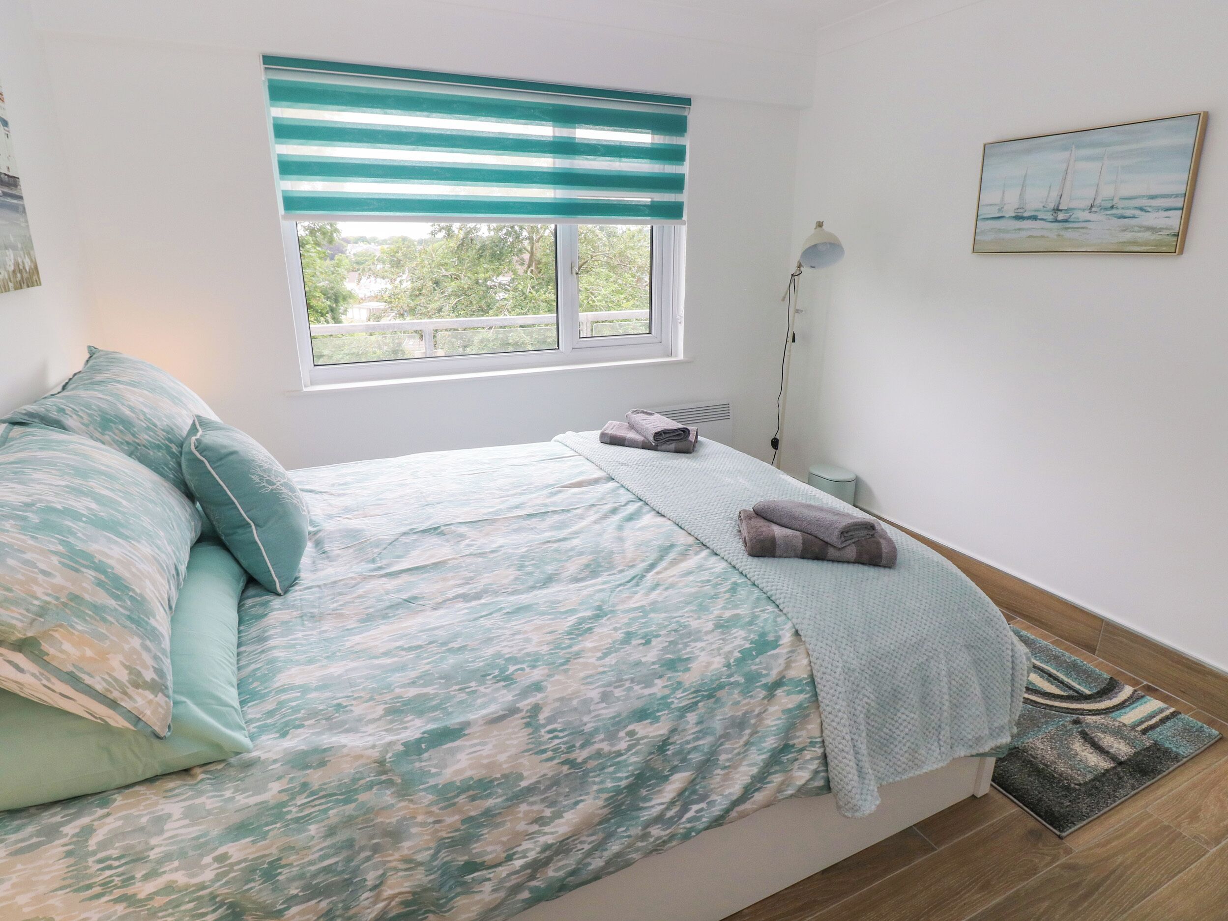 A bedroom with a bed, a window with a view, a painting on the wall, and a floor lamp.