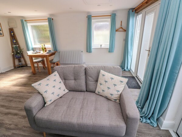 A cozy living room with a grey sofa, two matching pillows, a wooden dining table with chairs, and a window with light blue curtains. - Tower Cottage (Pembroke)