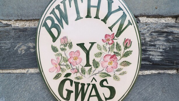 A circular sign with the text "BWTHYN Y GWAS" and floral illustrations. - Bwthyn y Gwas (Tywyn)