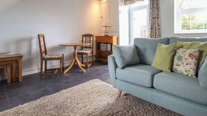 Ein gemütliches Wohnzimmer mit einem blauen Sofa, einem Holzesstisch mit Stühlen und einem großen Fenster mit floralem Vorhang.