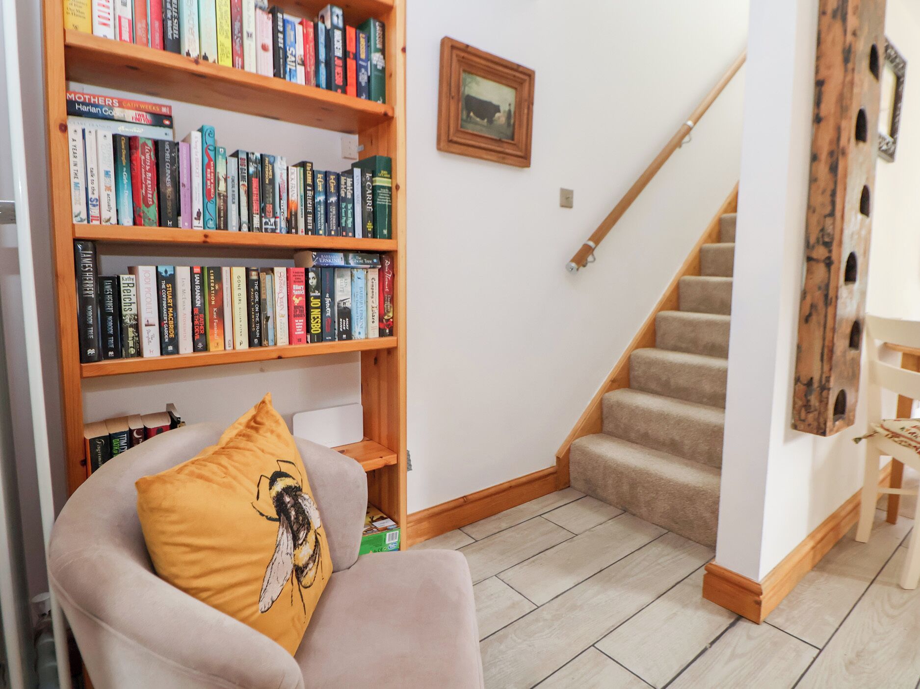 A living room with a bookshelf, a chair with a bee pillow, a framed picture on the wall, and a wooden staircase.
