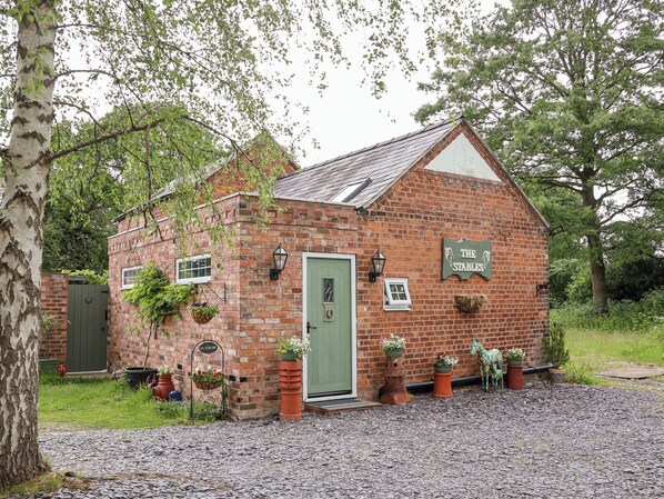 A brick building with a green door, labeled "The Stables," surrounded by greenery and potted plants. - The Stables (Chester)