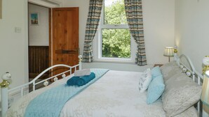 A bedroom with a bed, a window with curtains, a bedside lamp, and a door. - The Coach House (Dawlish)