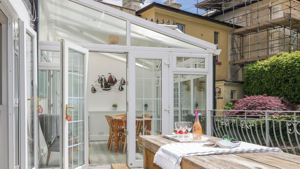 A covered patio area with a wooden bench and table, a glass door leading to an interior space with a dining table and chairs, and a view of a building with scaffolding. - The Coach House (Dawlish)