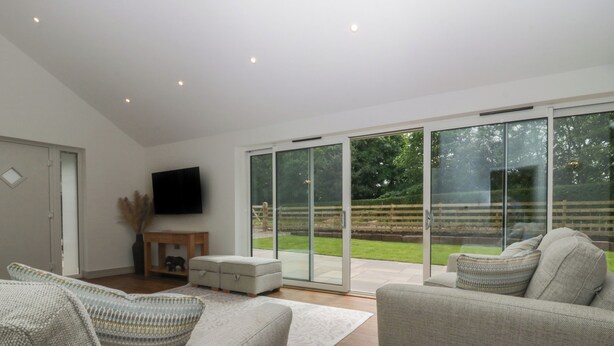 A modern living room with a large sliding glass door leading to a garden. There is a flat-screen TV mounted on the wall, a wooden coffee table, and two sofas. - Heather's Cottage (Carlisle)