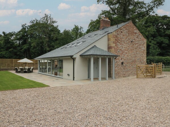 A stone building with a sloped roof, a covered patio area, and a gravel driveway. - Heather's Cottage (Carlisle)