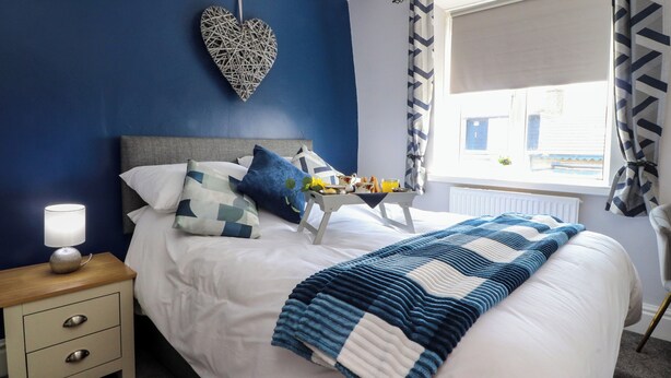 A neatly made bed with a blue and white striped blanket, a heart-shaped wall decoration, and a bedside table with a lamp. - Verdo House (Blackpool)