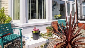 A house with a white exterior, a large window with vertical blinds, a green bench, and potted plants. - Verdo House (Blackpool)
