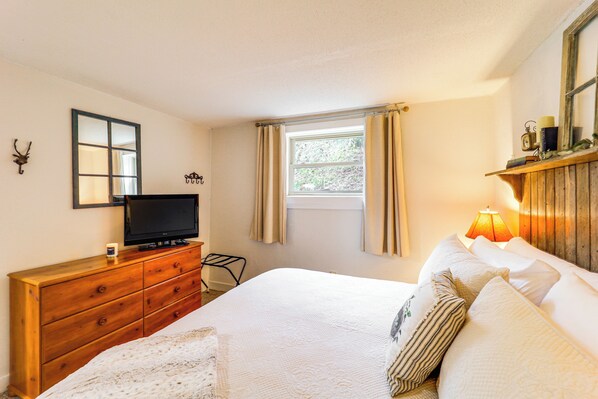 A bedroom with a wooden dresser, a television, a bed with white bedding, a window with curtains, and a wall-mounted shelf with decorative items. - Hike, Sip & Stay Awhile! Sugar Mountain Getaway (Banner Elk)