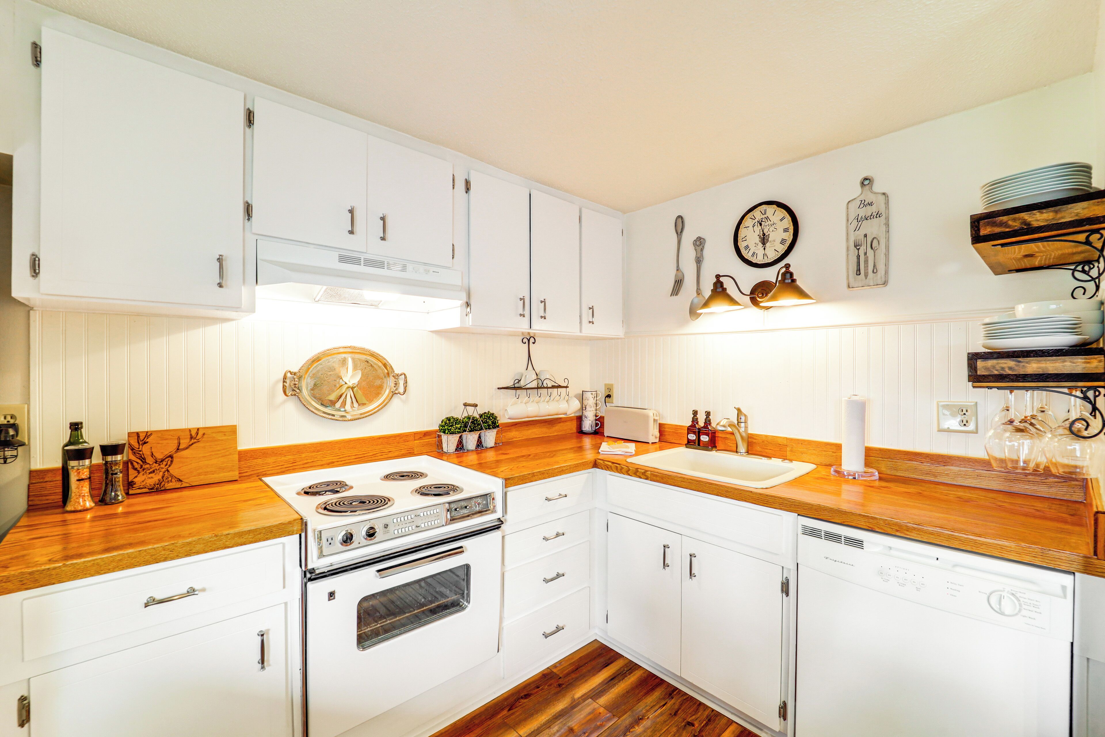 A kitchen with white cabinets, a stove, a sink, and a dishwasher. There are wall-mounted shelves with plates and a clock.