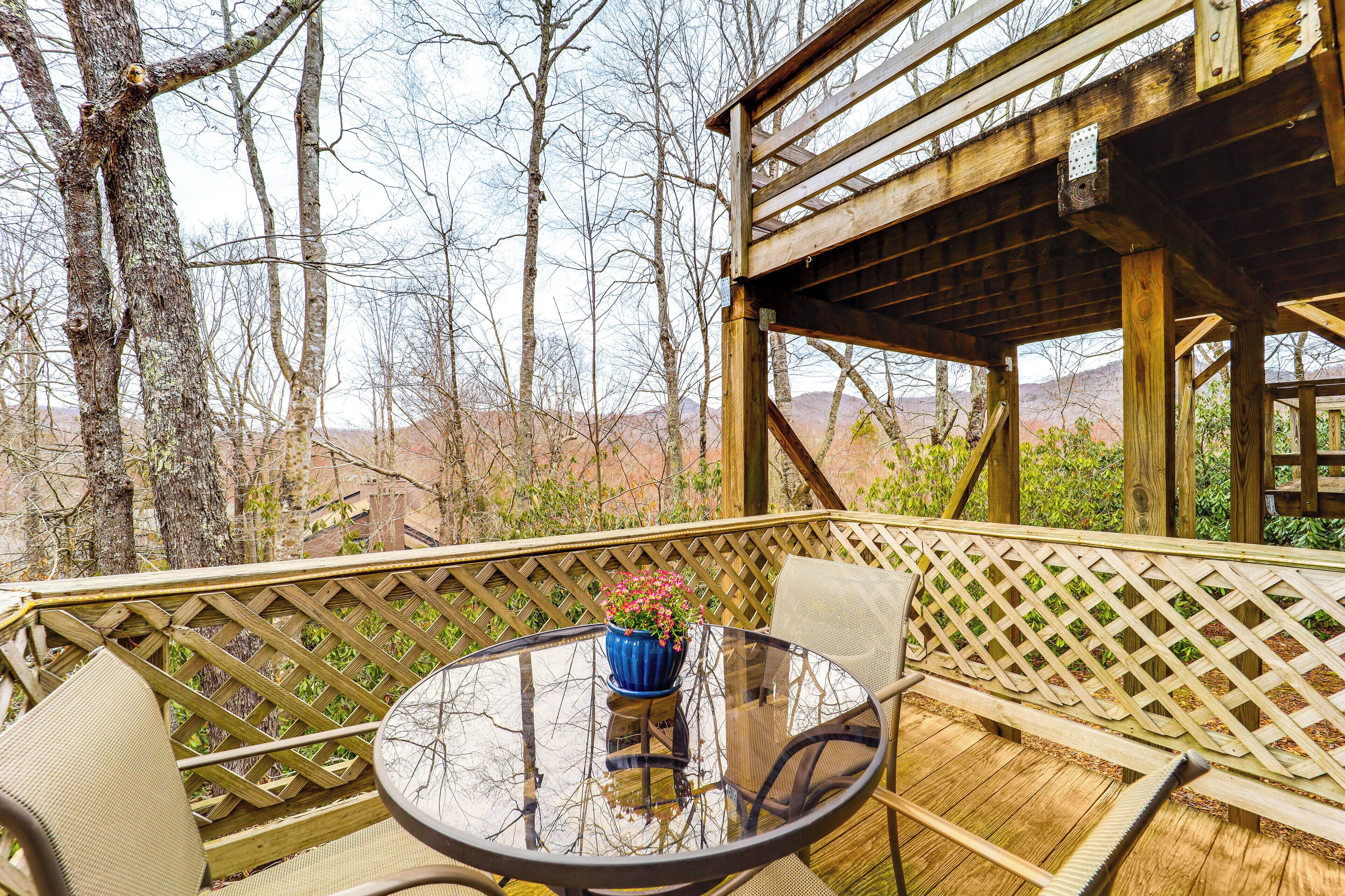 An outdoor wooden deck with a round glass table and chairs, surrounded by a wooden lattice fence, overlooking a mountainous landscape.