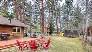 A wooden cabin with a deck, red Adirondack chairs, a grill, and a trampoline.