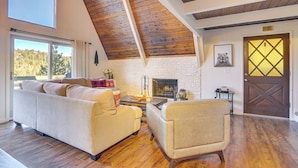 A cozy living room with a fireplace, wooden beams, and a large window overlooking a forest. - Family-friendly Lake Arrowhead Cabin w/ Game Room! (Lake Arrowhead)