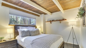 A bedroom with a wooden ceiling, a large window, a bedside table with a lamp, a bed with a white comforter and pillows, and a floor lamp. - Family-friendly Lake Arrowhead Cabin w/ Game Room! (Lake Arrowhead)