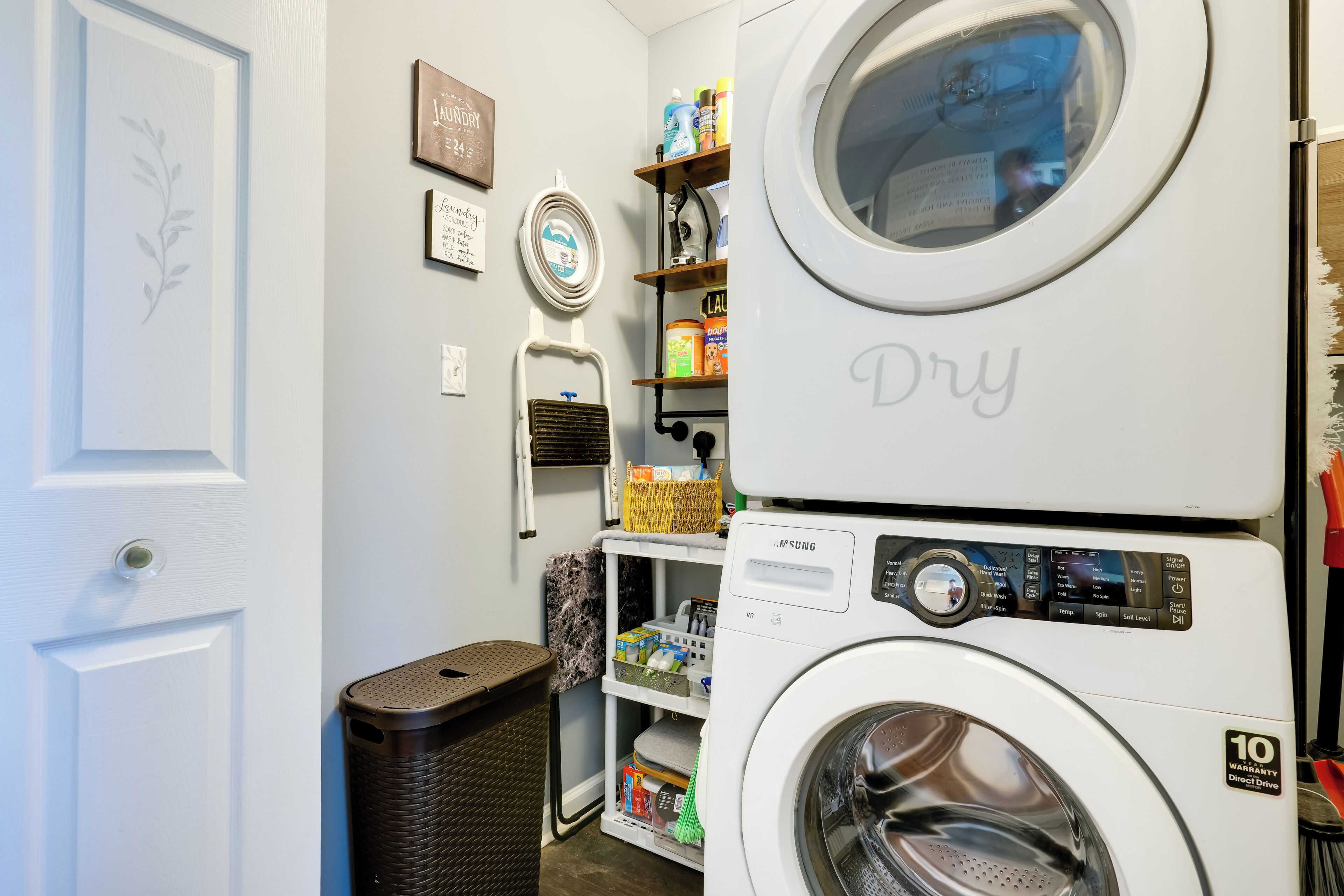 A laundry area with a Samsung washer and dryer, a trash bin, and shelves with laundry supplies.