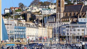 A harbor with boats, a church with a tower, and residential buildings in the background. - The Grottoes (Torquay)