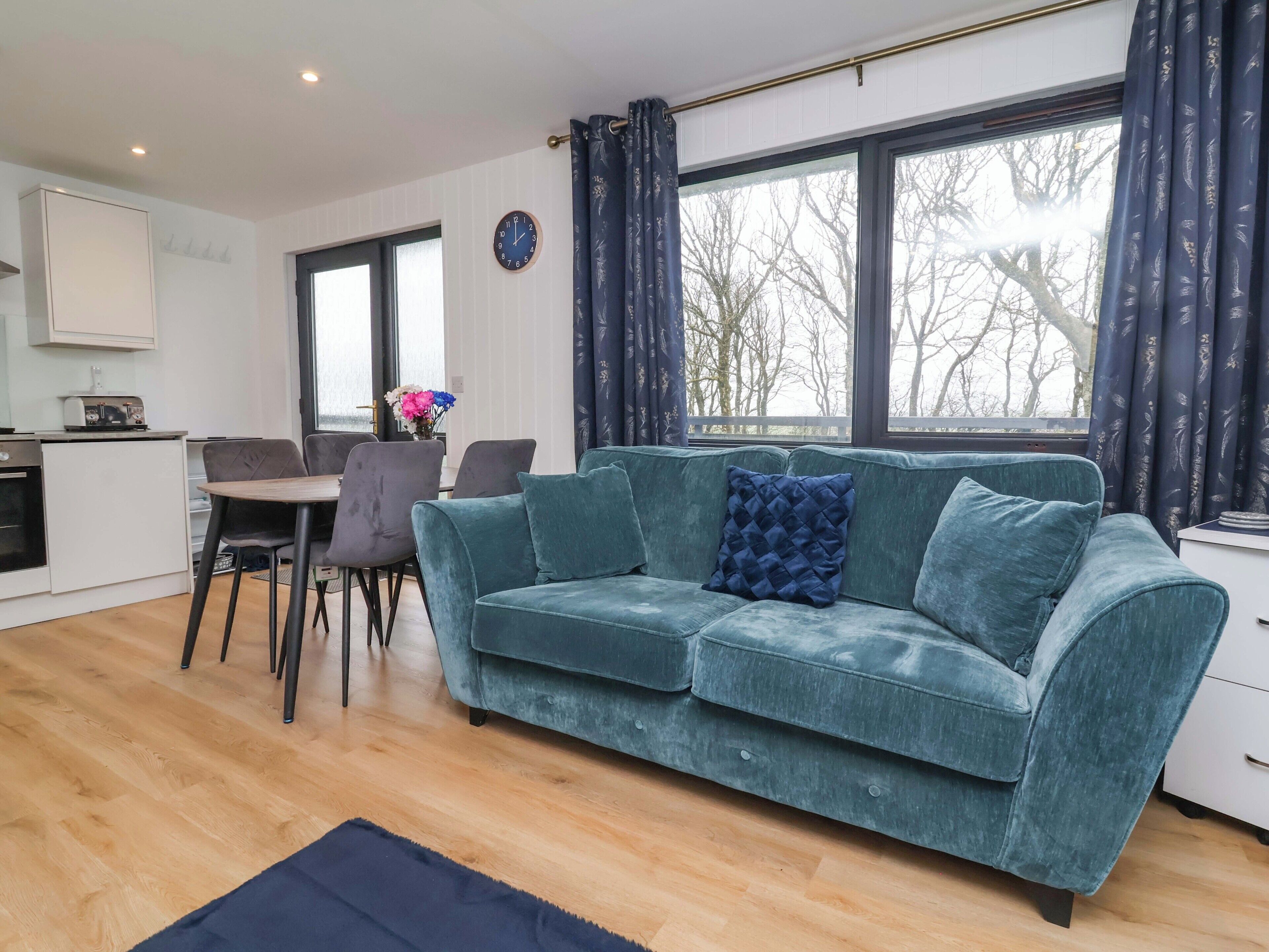 A living room with a sofa, a dining table with chairs, a kitchen area, and large windows with curtains.