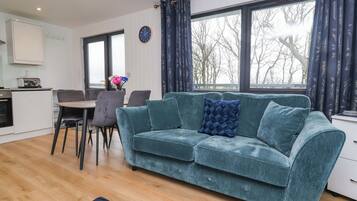 A living room with a sofa, a dining table with chairs, a kitchen area, and large windows with curtains.
