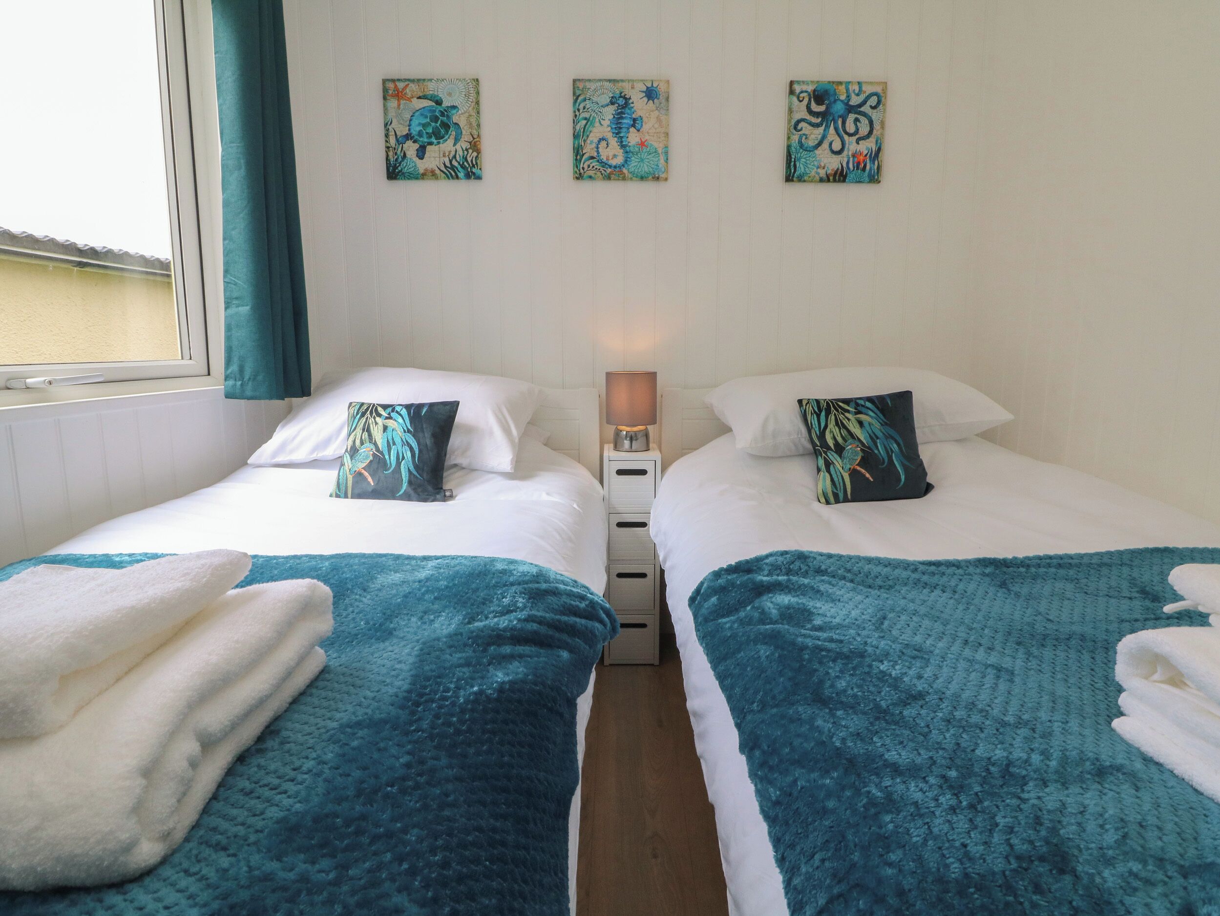 Una habitación de hotel con dos camas, cada una con una manta color turquesa y almohadas blancas, una mesita de noche con una lámpara, y tres obras de arte colgadas en la pared que representan criaturas marinas.