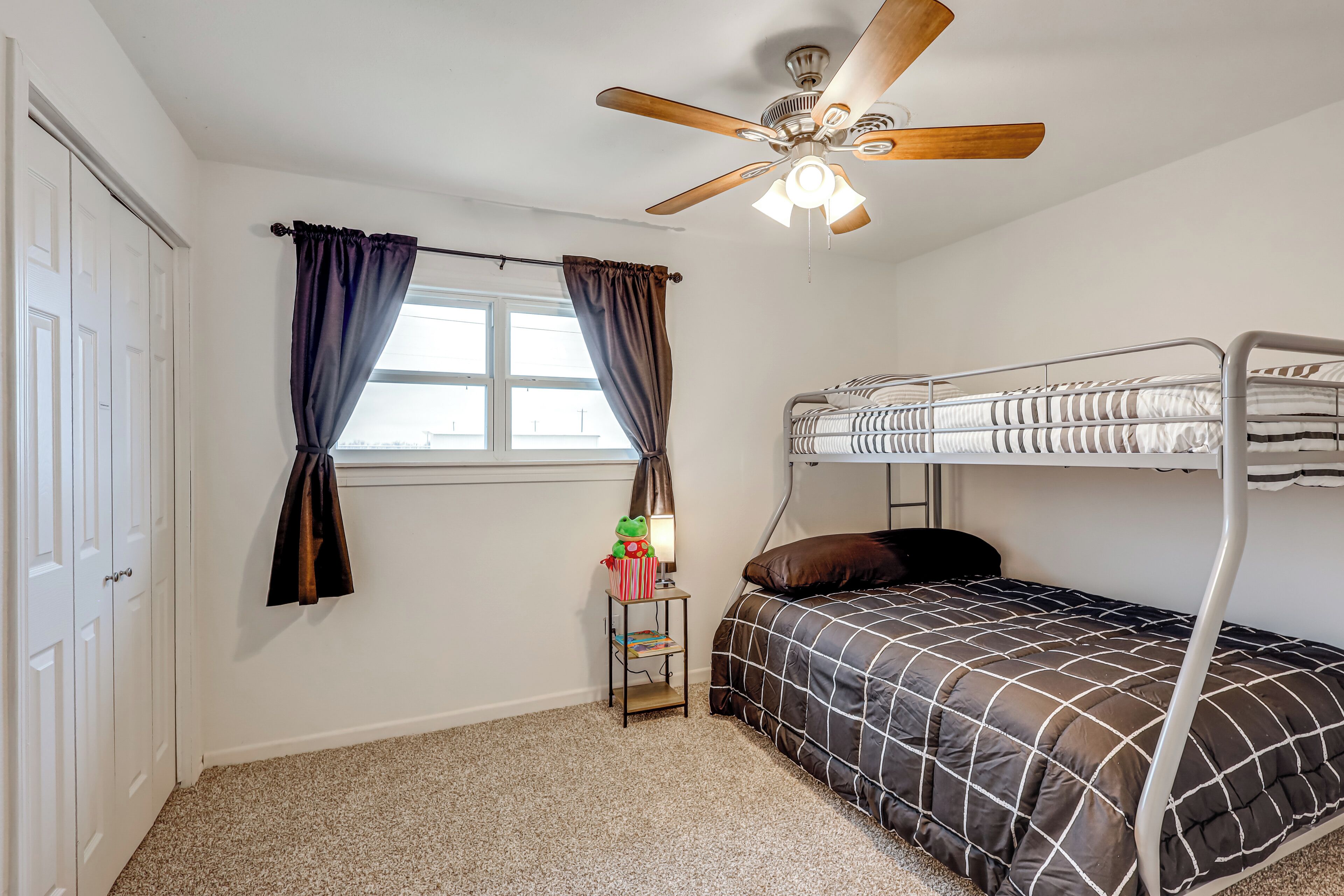 A bedroom with a bunk bed, a ceiling fan, a window with curtains, and a small table with a lamp and books.