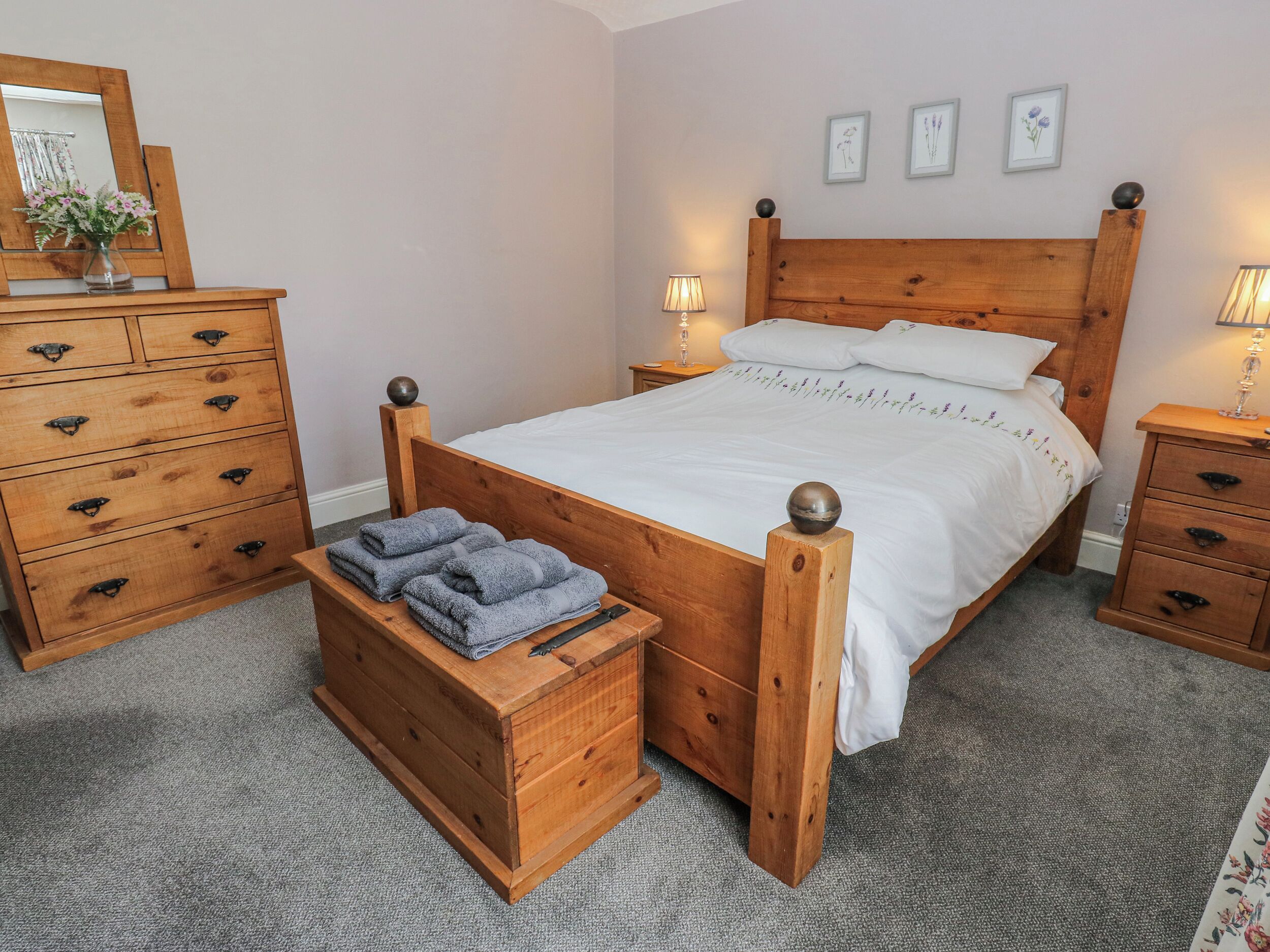 A wooden four-poster bed with a white bedspread, two bedside lamps, a wooden nightstand, a wooden dresser with multiple drawers, and a small wooden chest with folded towels.