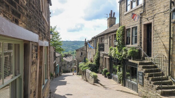 A narrow cobblestone street lined with historic stone buildings, including a pub with outdoor seating and a staircase leading to an upper floor. - Penny Cottage (Keighley)