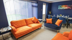 A room with a blue wall, a large window with blinds, a blue sofa, an orange sofa, a small table, and a dining area with blue chairs and a table set for a meal.