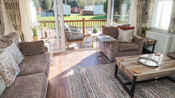 A spacious living area with a wooden floor, a sofa, a coffee table, and a sliding glass door leading to an outdoor deck. - Acorn Lodge (Morpeth)