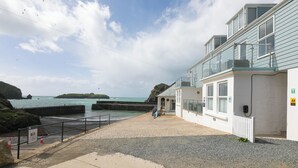 A coastal building with a balcony overlooking the sea, a paved walkway, and a rocky shoreline. - Demelza (Helston)