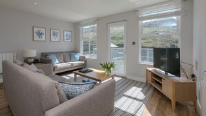 A modern living room with a sofa, coffee table, and TV stand. There are large windows with a view of hills. - Demelza (Helston)