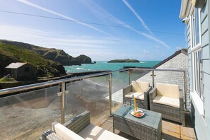A balcony with wicker furniture, a glass railing, and a view of the sea and cliffs. - Demelza (Helston)