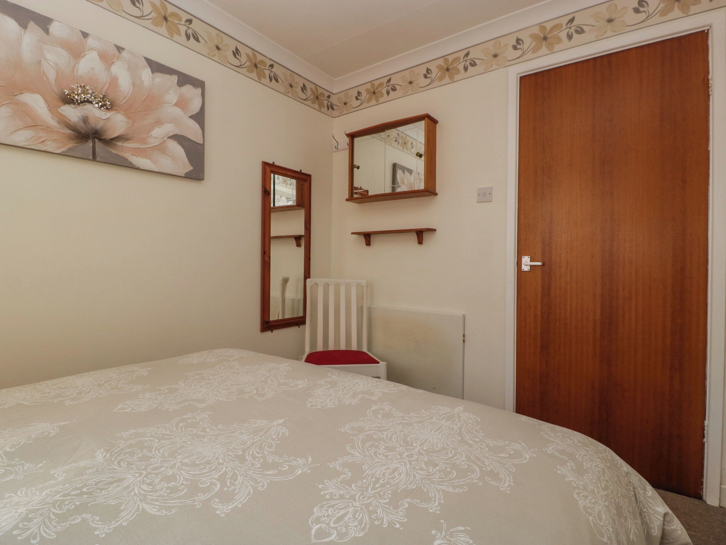 A bedroom with a bed, a wooden door, a radiator, a mirror, and a floral wall decoration.
