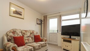 A living room with a floral sofa, a television on a wooden stand, a radiator, and a window with curtains. - Blorenge (Abergavenny)