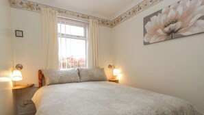 A bedroom with a large bed, two bedside lamps, a window with curtains, and a floral painting on the wall. - Blorenge (Abergavenny)