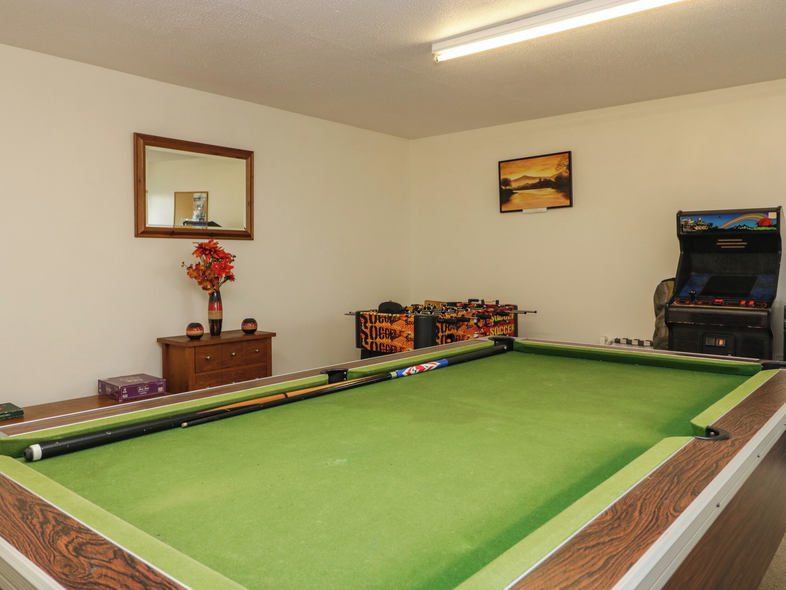 A pool table with a green felt surface, a wooden cabinet with decorative items, a framed mirror, and a wall-mounted painting.
