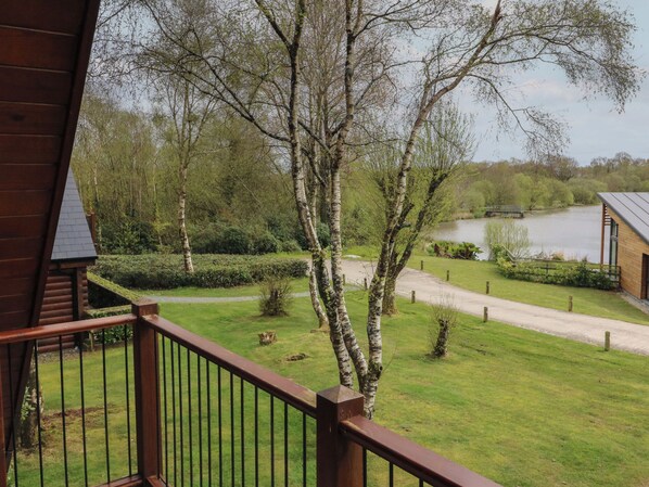 A view from a wooden balcony overlooking a grassy area with trees and a lake. - Cedar (Winkleigh)