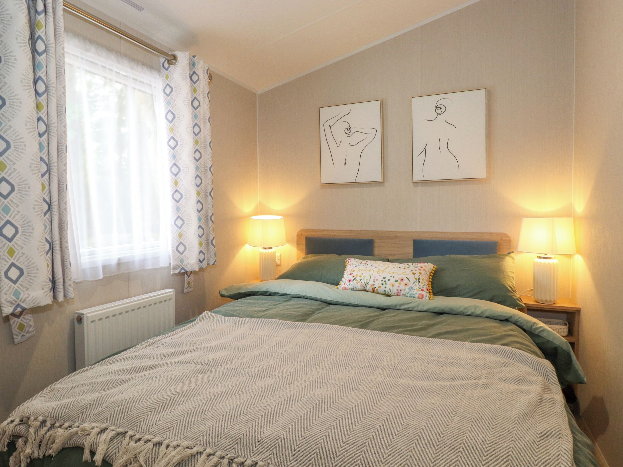 Une chambre à coucher comprenant un lit, deux lampes fixées au mur, une fenêtre avec des rideaux à motifs et deux œuvres d’art mural.