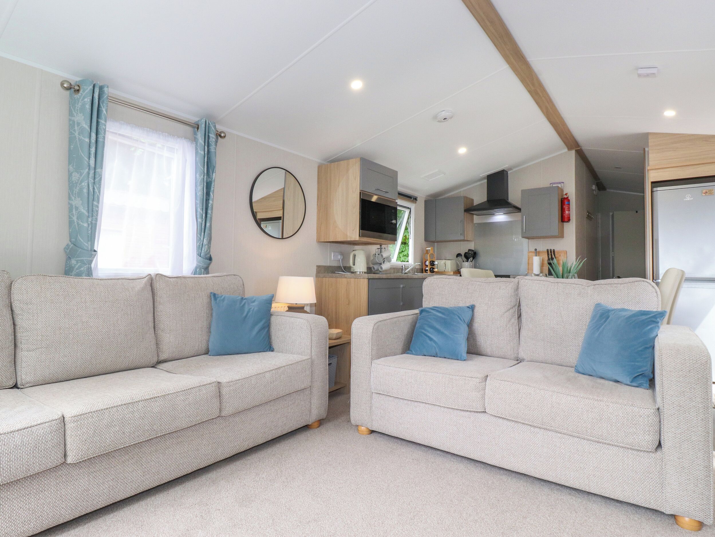 A modern living room with two grey sofas, a round mirror, and a kitchen area with wooden cabinets and a built-in oven.
