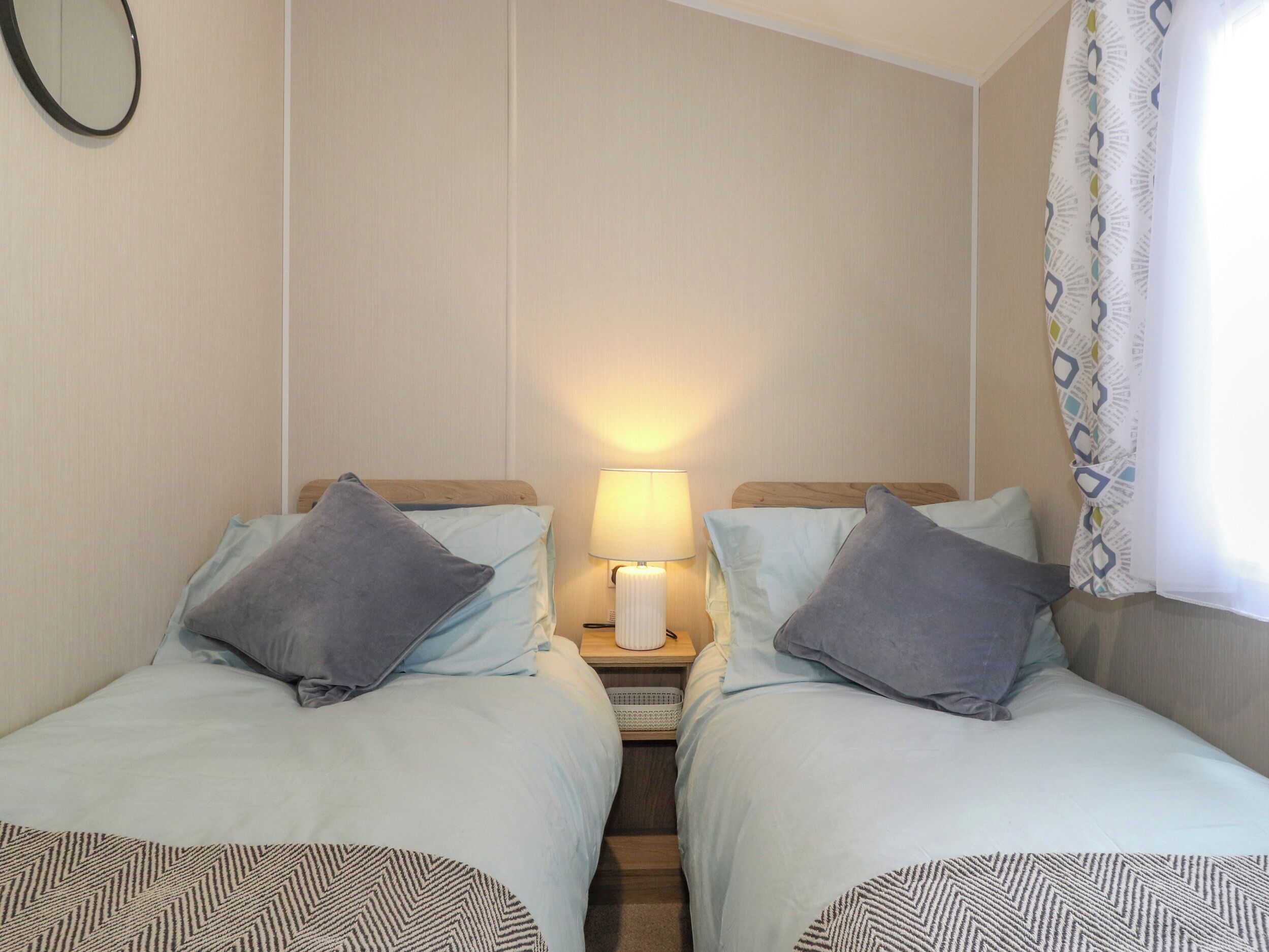 A twin room with two beds, each with a pillow and a patterned throw. There is a nightstand with a lamp between the beds.
