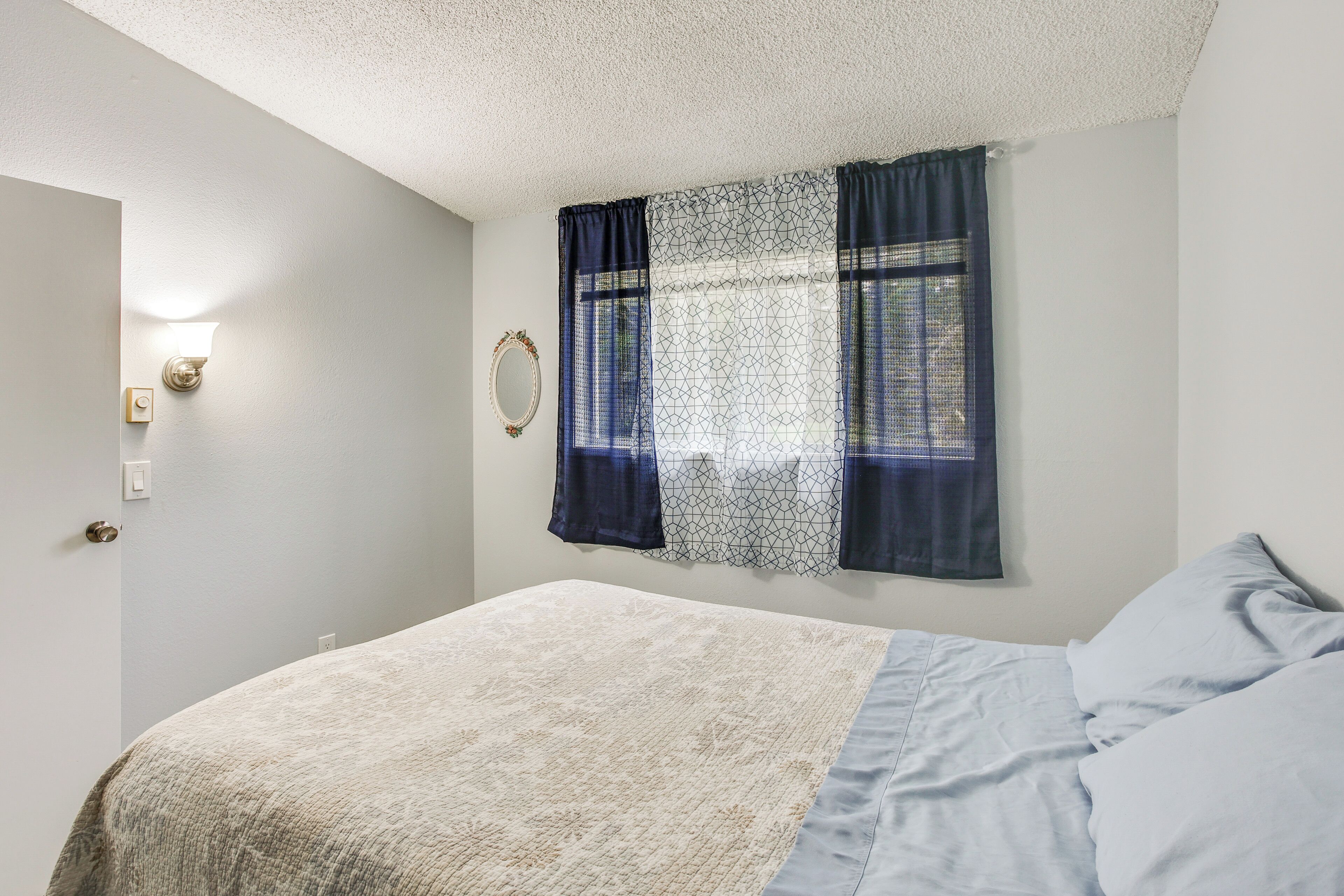 Quarto com cama, janela com cortinas estampadas, luminária fixada na parede e porta com puxador.