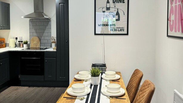 A modern kitchen with dark cabinetry, a stainless steel range hood, and a dining area set for a meal with white plates and bowls. - Modern 1-bed Apartment - Sleeps 4 & Central Links (Birkenhead)