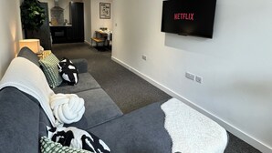 A modern living room with a grey sofa, a white throw blanket, and a flat-screen TV mounted on the wall displaying "NETFLIX". - Modern 1-bed Apartment - Sleeps 4 & Central Links (Birkenhead)