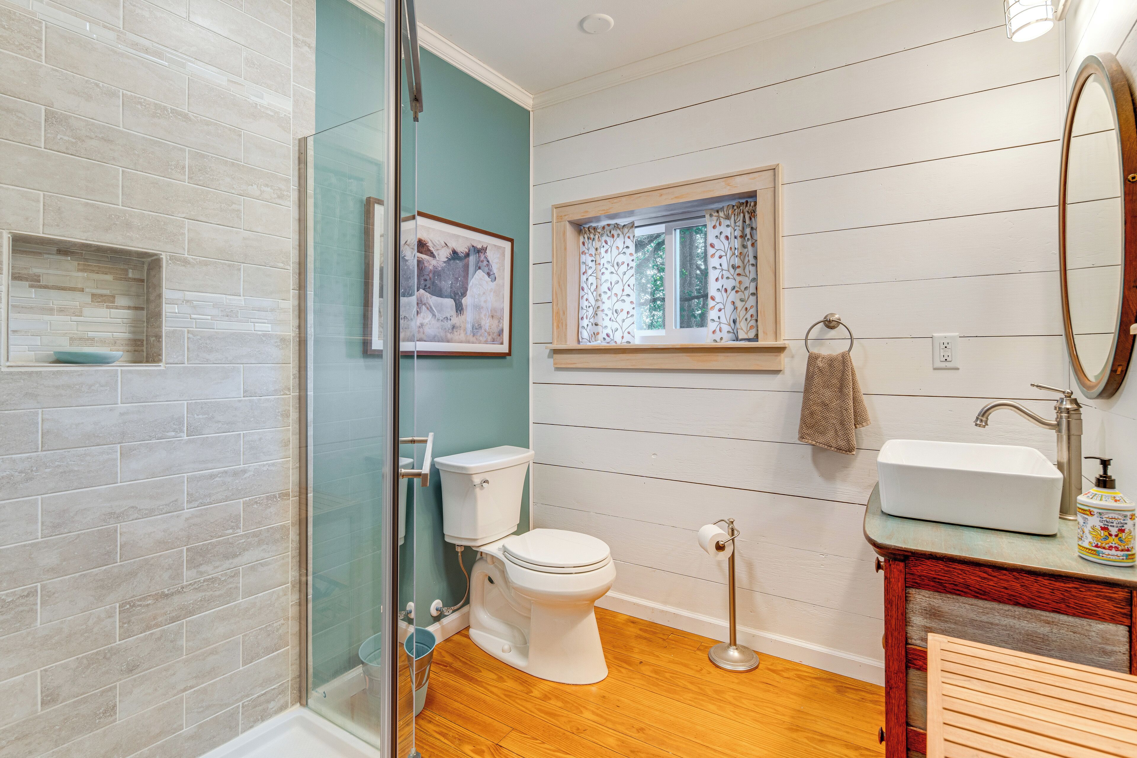A bathroom with a glass shower enclosure, a toilet, a sink with a mirror, and a window with a curtain.