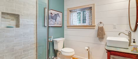 A bathroom with a glass shower enclosure, a toilet, a sink with a mirror, and a window with a curtain.