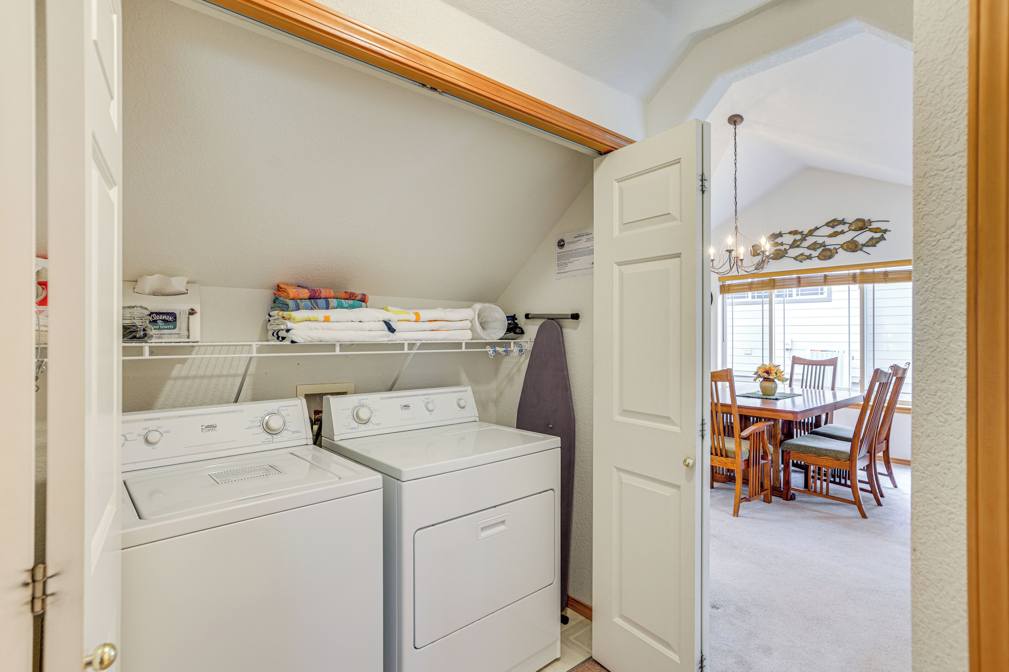 A laundry room with two stacked washers and dryers, a drying rack, and a dining area with a table and chairs visible through an open door.