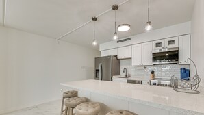 A modern kitchen with white cabinetry, a stainless steel refrigerator, a built-in oven, and a marble backsplash. There are four bar stools with cushioned seats. - Dogs Welcome: Beachfront South Padre Island Condo (South Padre Island)