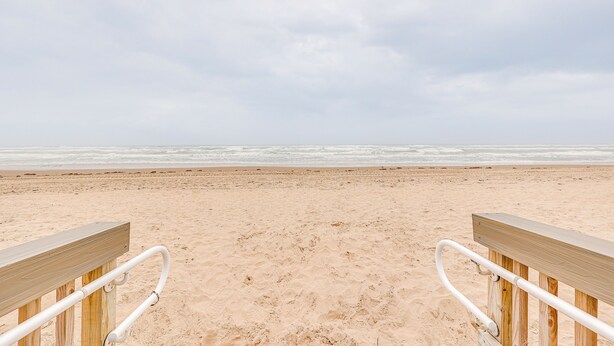 A wooden bench with metal handrails on a sandy beach with the ocean in the background. - Dogs Welcome: Beachfront South Padre Island Condo (South Padre Island)